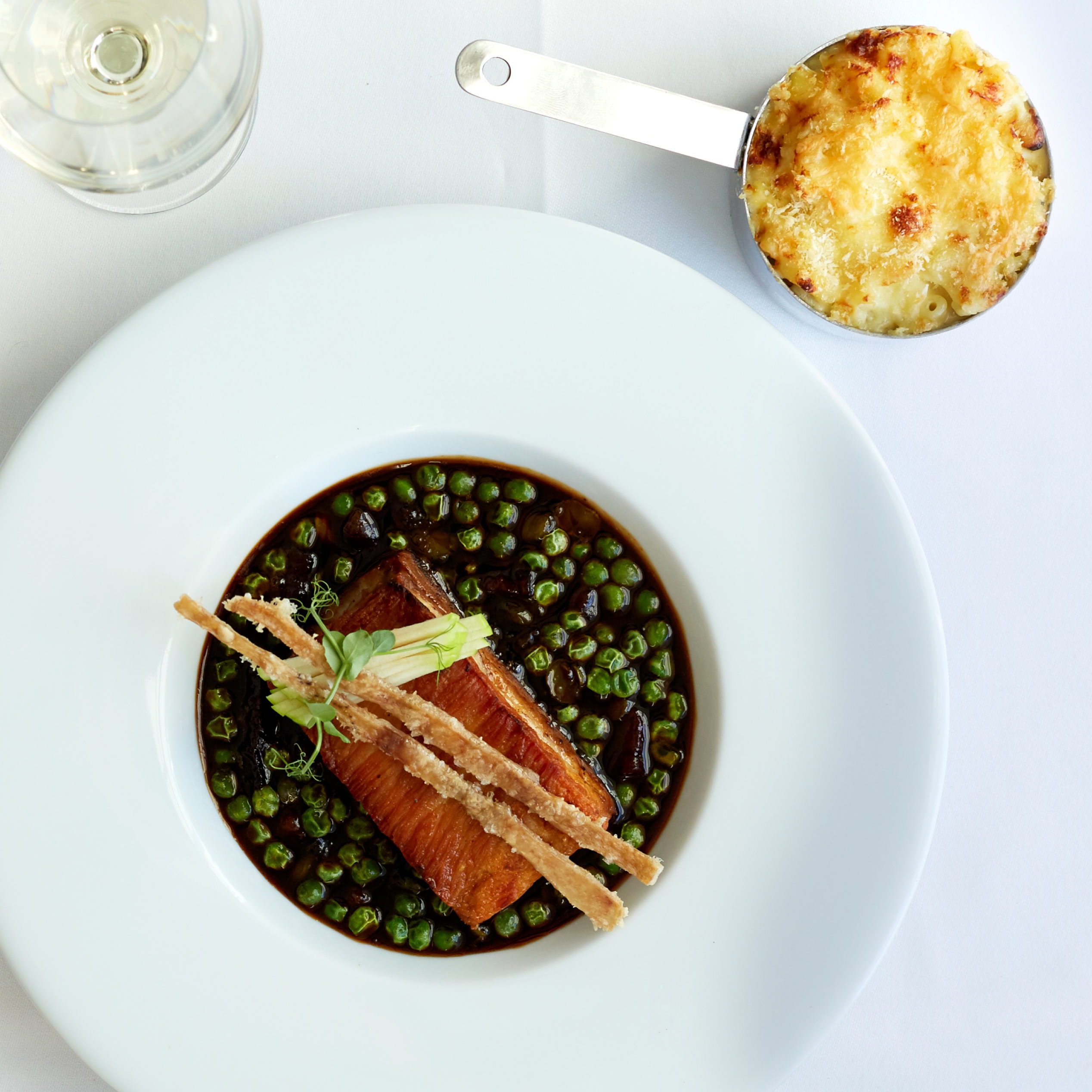 Overhead shot of plated main course in dark sauce with peas, with a baked side dish in a metal pan