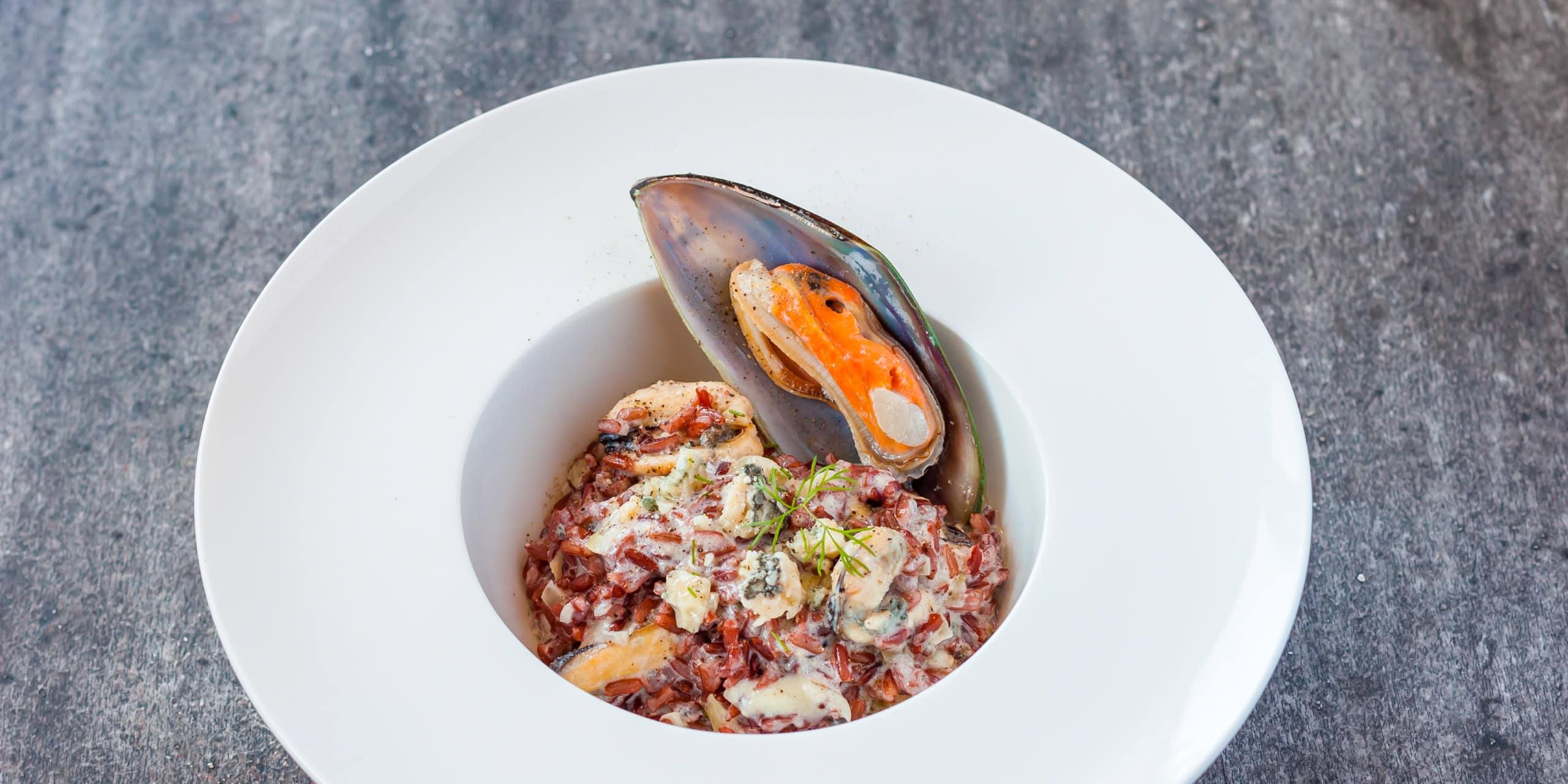 Seafood risotto in a white bowl topped with a mussel, photographed on a textured surface