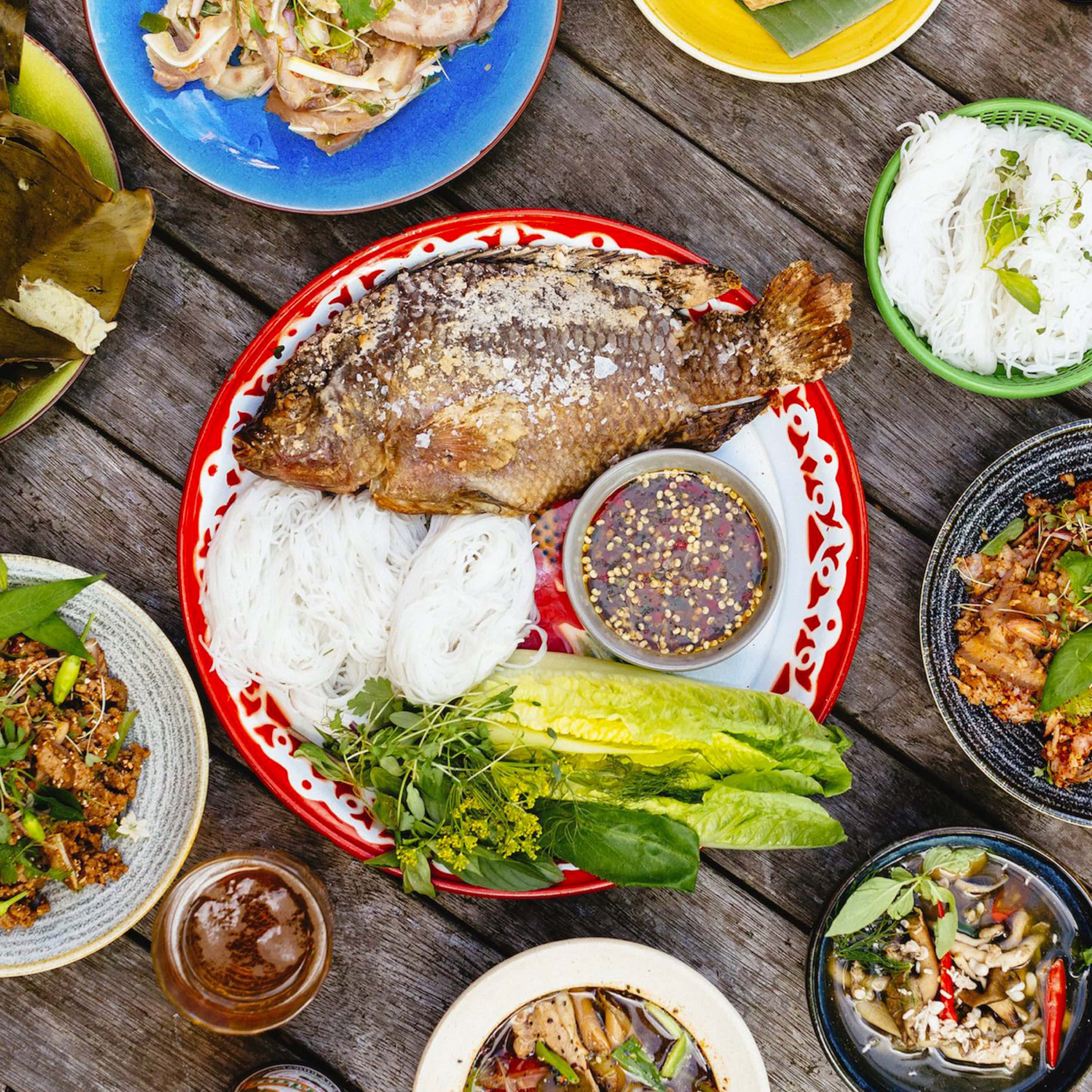 Overhead spread of Lao dishes with whole fried fish, noodles, herbs and dipping sauce on a wooden table