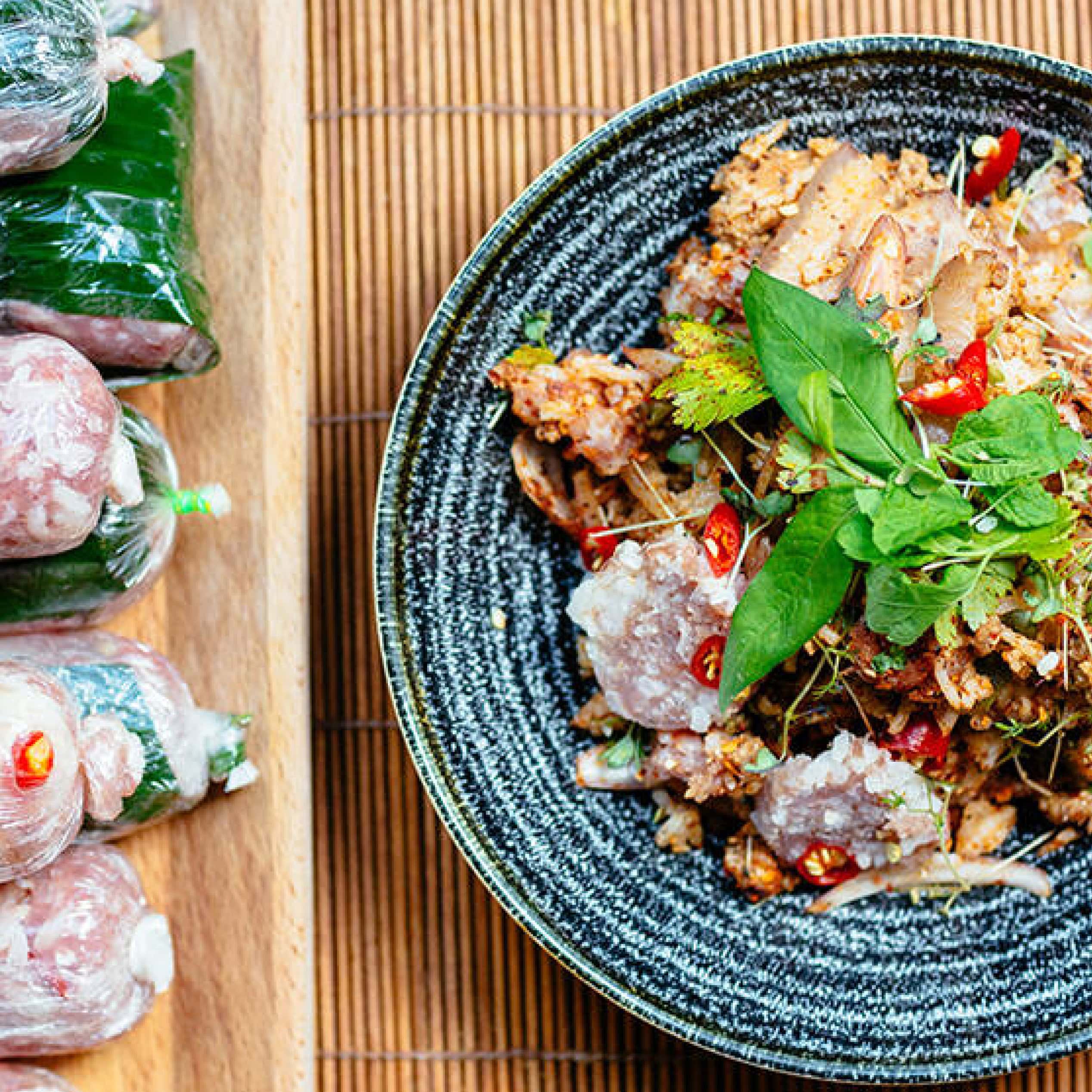 Close-up of a Lao spicy salad topped with herbs and chilli, beside wrapped ingredients on a board