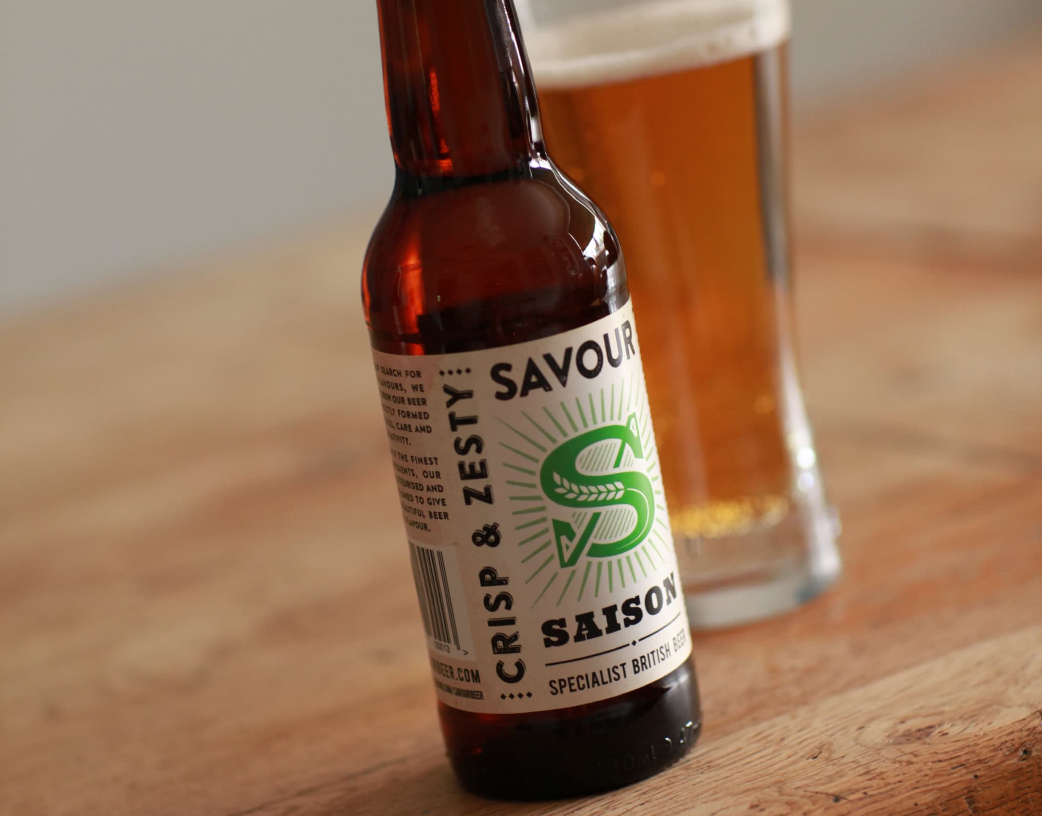 Savour Saison beer bottle on a wooden table with a glass of beer in the background