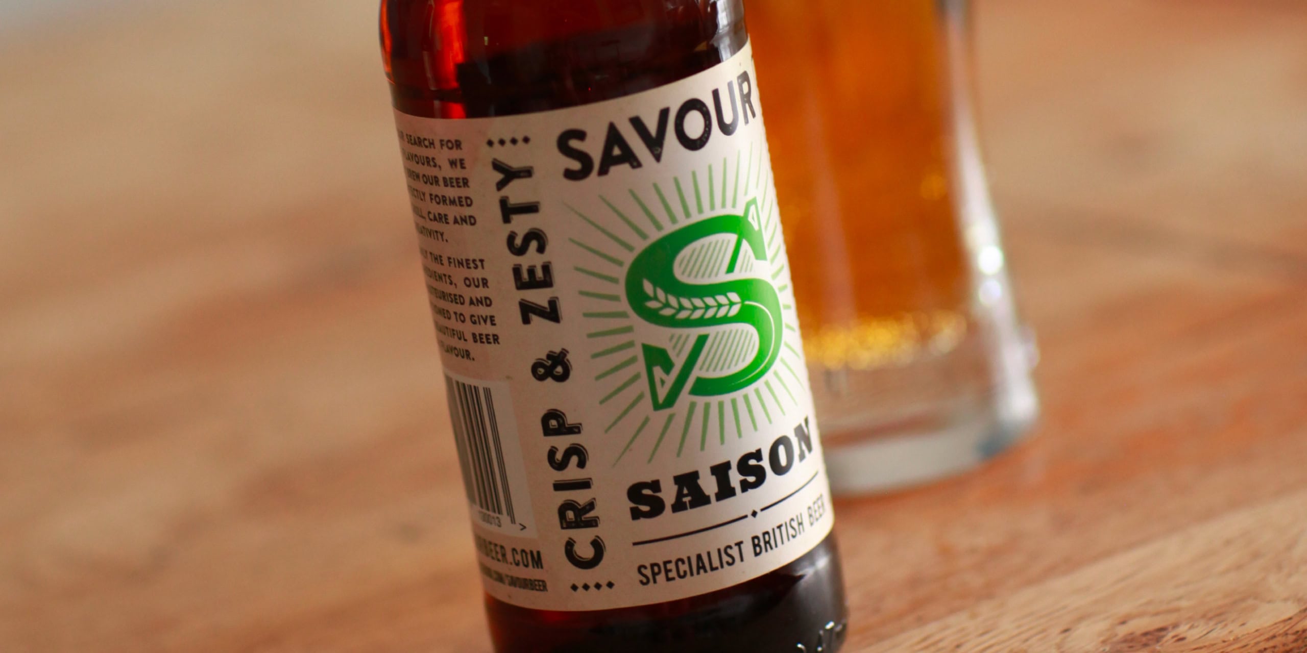 Savour Saison bottle with green “S” label beside a glass of beer on a wooden table