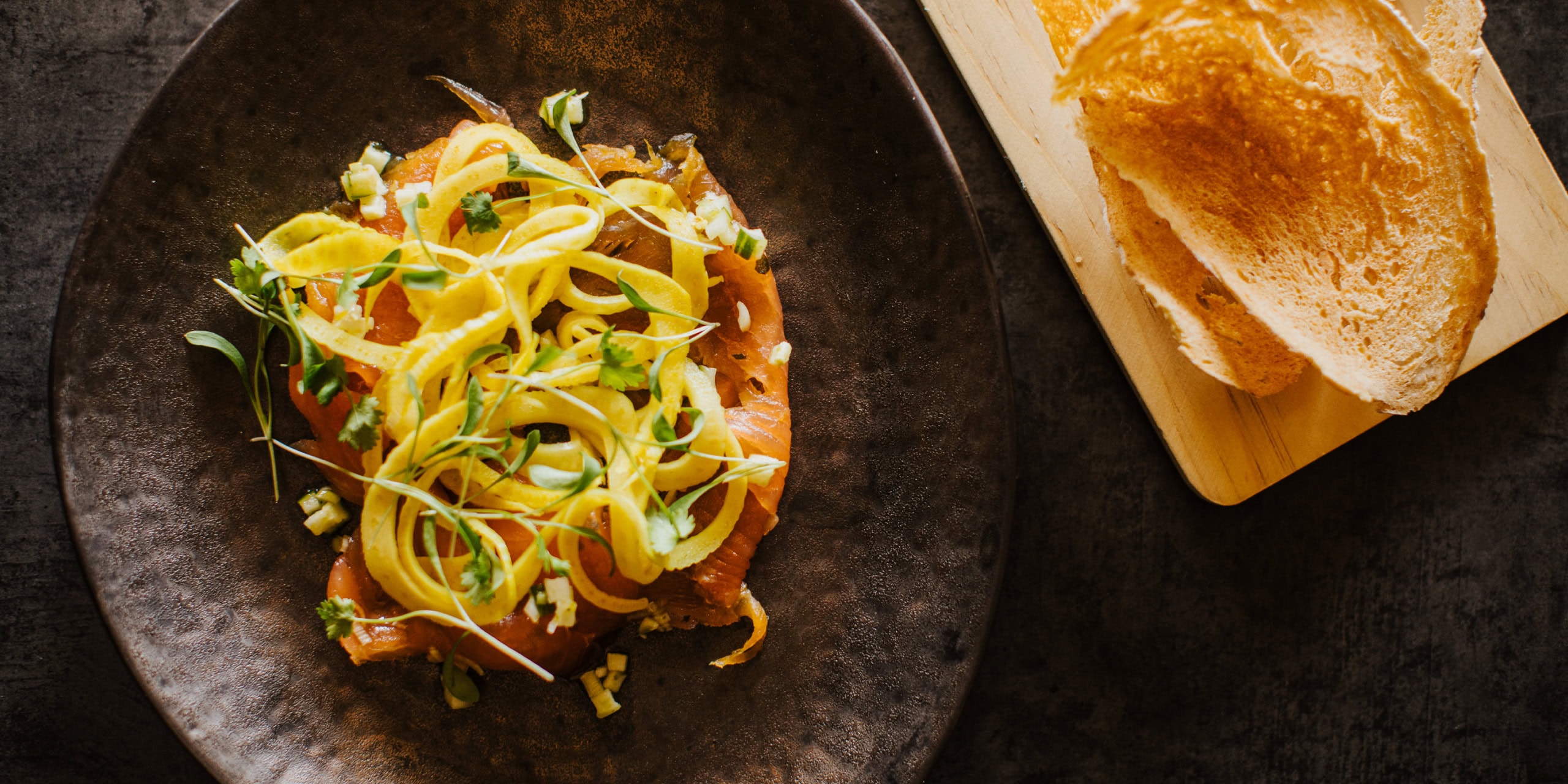 Overhead shot of smoked salmon topped with ribboned courgette, microgreens and diced garnish on a dark plate, with toast and spread on a wooden board.